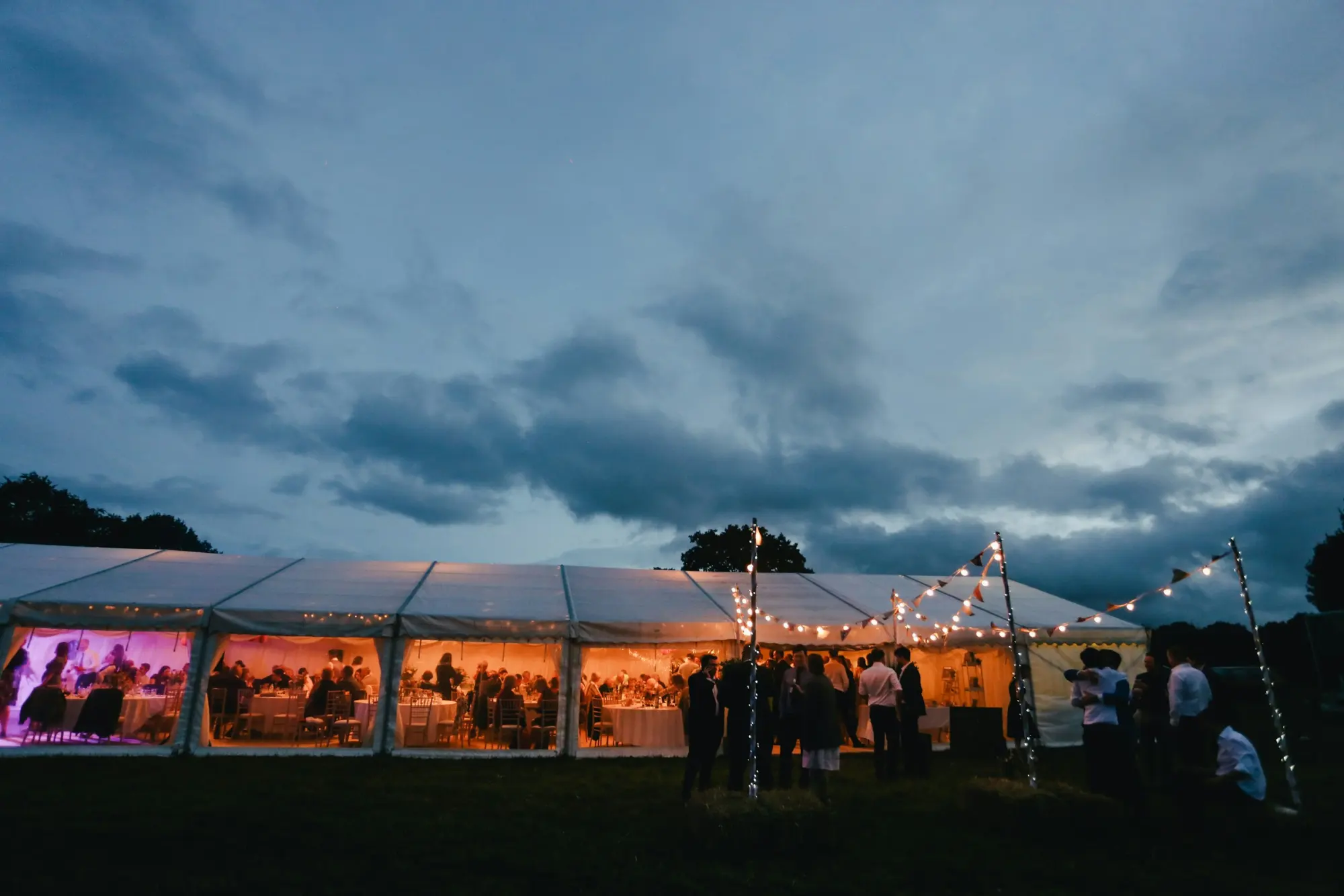 Sample photo: a long white frame tent lit up at dusk, guests at round tables inside, string lights on poles out front